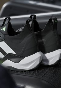 Paire de chaussures de sport noires et blanches avec des détails verts sur le logo, placées sur une surface noire texturée, mettant en avant leurs semelles épaisses et anguleuses.