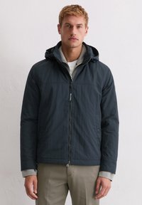 Granatowy płaszcz z kapturem, z przednim zamkiem błyskawicznym, elastycznymi mankietami i teksturowanym wykończeniem. Warstwowo założony na szarą bluzę.