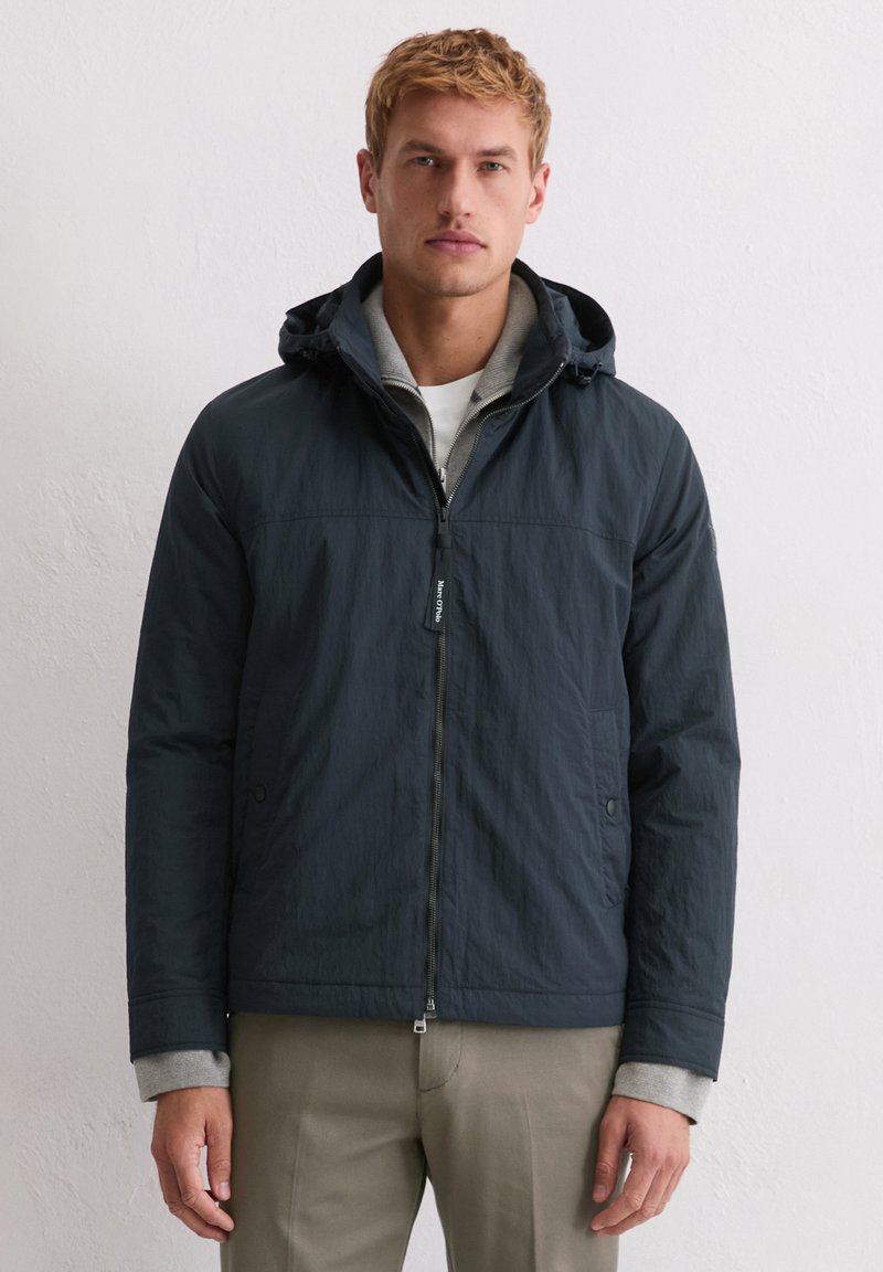 Granatowy płaszcz z kapturem, z przednim zamkiem błyskawicznym, elastycznymi mankietami i teksturowanym wykończeniem. Warstwowo założony na szarą bluzę.