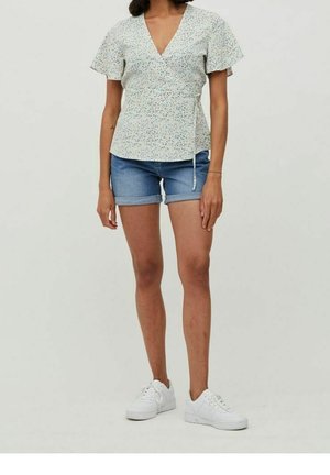 Nainen, joka on pukeutunut lyhythihaiseen kukalliseen kietaisupaitaan, farkkushortseihin ja valkoisiin tennareihin, seisoo yksinkertaisen vaalean taustan edessä.