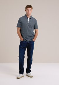 Mladic stoji uz neutralnu pozadinu, odjeven u tamnoplavu polo majicu, plave traperice i bijele tenisice, s rukama u džepovima.