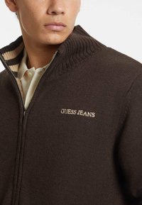 Barna kötött, cipzáras pulóver bordás gallérral, krémcsíkos belső résszel, a mellkason világos bézs színű "GUESS JEANS" hímzéssel.