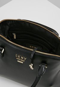 Černá kožená kabelka DKNY New York se zlatým zipem otevřená, ukazující vnitřní zipovou kapsu a značkovou podšívku.