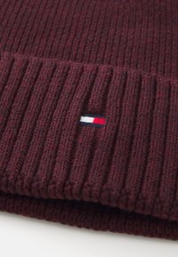 Burgundowa dzianinowa czapka z prążkowanym wzorem, z małym wyszywanym logo w czerwono-biało-niebieskich akcentach. Wykonana z miękkiej wełniano-polyesterowej mieszanki.
