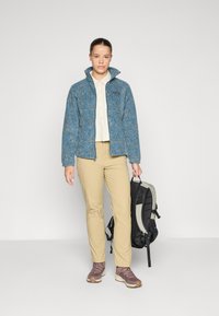 Blau gemusterte Jacke mit hohem Kragen, beiges Hemd, beige Hose und dunkler Rucksack. Braune Wanderschuhe mit Schnürsenkeln. Einfaches, funktionales Design.