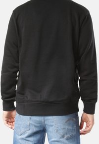 Svart sweatshirt med långa ärmar, ribbade muddar och fåll. Materialet verkar mjukt; baksidans design är enkel, utan synliga logotyper eller mönster.