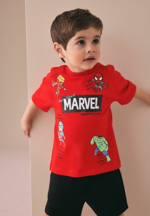 Egy fiatal fiú piros Marvel pólót visel, amelyen Iron Man, Pókember, Amerika Kapitány és Hulk illusztrációk láthatók, bézs fal előtt áll.