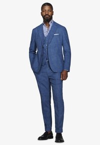 Uomo che indossa un abito blu a tre pezzi dalla trama strutturata con camicia a righe e scarpe eleganti nere, in piedi con le mani nelle tasche.