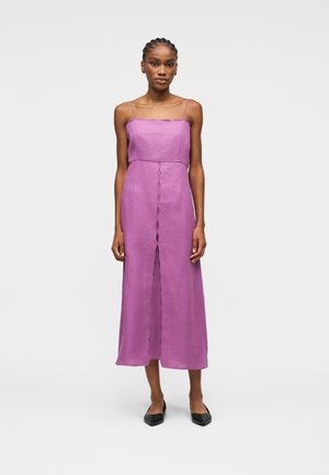 Mujer joven con el cabello trenzado, vestida con un vestido midi morado sin mangas con bordes festoneados y zapatos planos negros de punta, de pie, erguida.