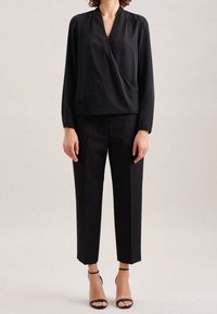 Blusa nera drappeggiata con maniche lunghe, abbinata a pantaloni neri a vita alta. Presenta una texture leggera e liscia e tacchi con cinturino alla caviglia.