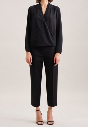 Blusa nera drappeggiata con maniche lunghe, abbinata a pantaloni neri a vita alta. Presenta una texture leggera e liscia e tacchi con cinturino alla caviglia.