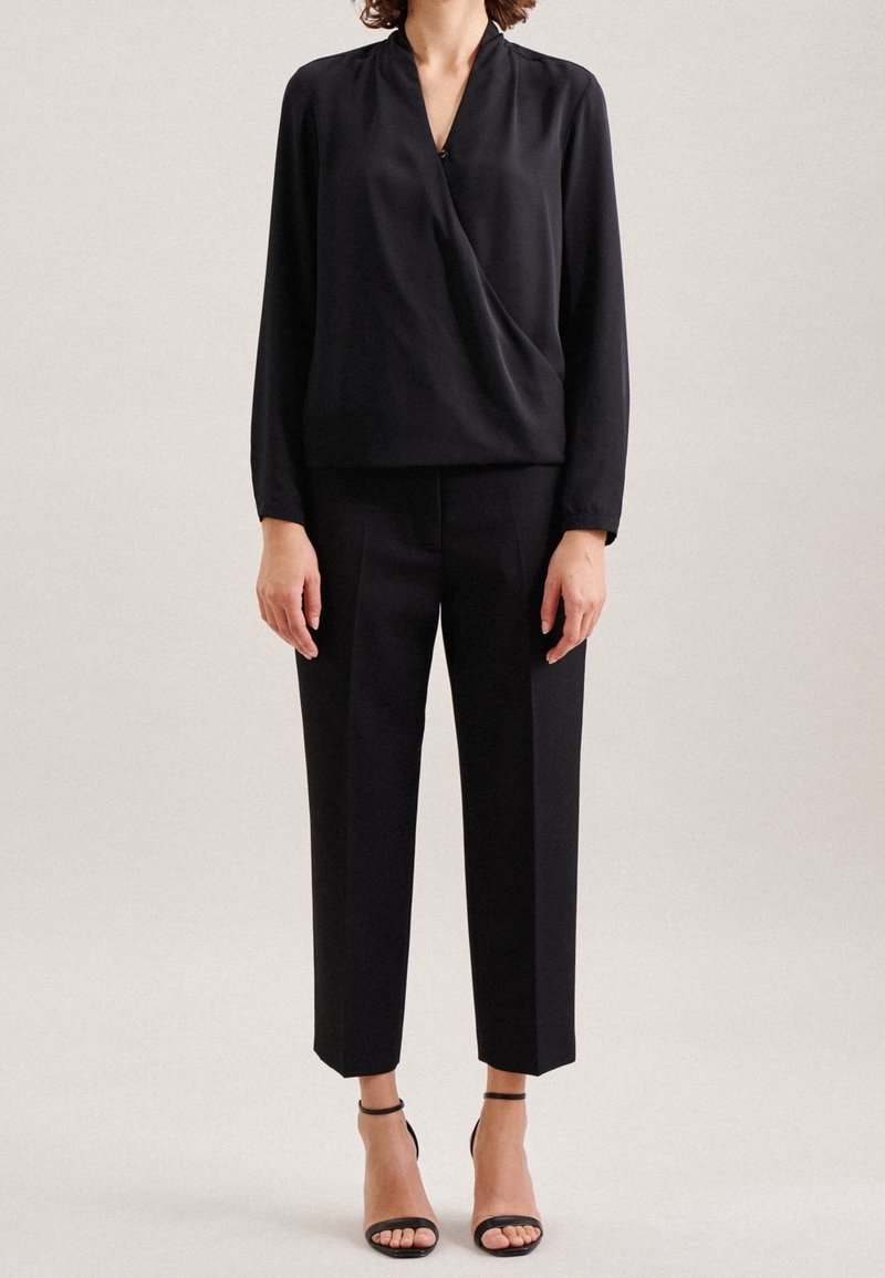 Blusa nera drappeggiata con maniche lunghe, abbinata a pantaloni neri a vita alta. Presenta una texture leggera e liscia e tacchi con cinturino alla caviglia.