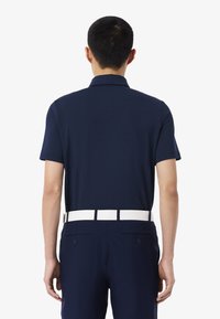 Polo shirt bleu marine à manches courtes, design col. La vue arrière montre une ceinture blanche et un pantalon ajusté avec deux poches arrière. Texture lisse.