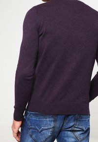 Pull violet foncé à col rond, manches longues et poignets côtelés. Porté avec un jean bleu arborant un subtil motif en denim et des détails de couture.