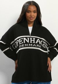 Pull en tricot noir avec un col zippé, arborant "COPENHAGUE DANEMARK" en lettres blanches sur le devant. Coupe ample avec des poignets côtelés.