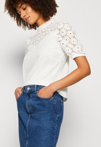 Hvid kortærmet top med en blonderede panel med cirkulære øjenlågsmønstre og en flæsekrave, parret med en blå denimnederdel.