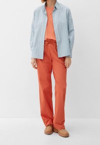 Lyseblå stribet skjorte over en orange t-shirt, parret med løstsiddende orange bukser og brune sko. Minimale detaljer, afslappet stil.