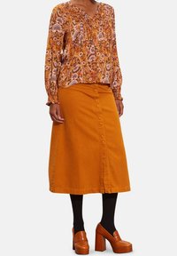 Blouse fluide et à motifs dans des tons chauds, associée à une jupe en denim, longueur genou, boutonnée et de couleur moutarde. Accent : escarpins à plateforme épais.