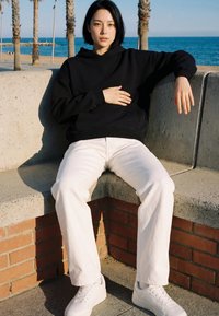 Personne en sweat à capuche noir et pantalon blanc assise sur un banc en béton au bord de la mer, palmiers en arrière-plan, une main sur la poitrine et l'autre reposant sur le banc.