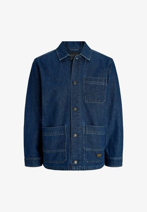 Junge Person steht und lächelt, trägt eine blaue Jeansjacke, ein grünes Poloshirt, braune Hose und beige Schuhe vor weißem Hintergrund.