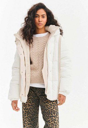 Biała pikowana kurtka z beżowymi akcentami, sweter o prążkowanej fakturze oraz spodnie w panterkę. Kurtka ma wysoki kołnierz i zatrzaski.