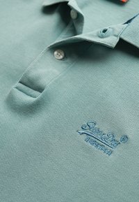 Polo shirt azzurro chiaro in tessuto testurizzato, con due bottoni al colletto e un logo ricamato di un blu più scuro.