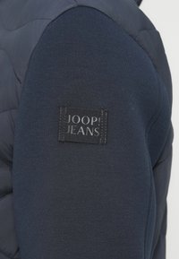 Σκούφο γιλέκο ναυτικού μπλε με ομαλό ύφασμα και υφασμένο σχέδιο. Διαθέτει μαύρη ετικέτα με το «JOOP! JEANS» κεντημένο σε περίοπτη θέση.