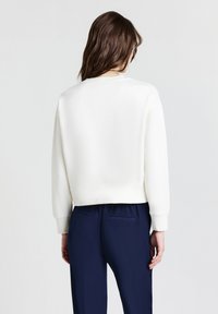 Una persona con capelli castani indossa una maglietta bianca a maniche lunghe e pantaloni blu scuro, in piedi con le spalle rivolte verso uno sfondo chiaro e uniforme.