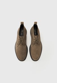 Zapatos de ante marrón con una textura suave, punta redonda, diseño con cordones y suela de goma negra contrastante. Etiqueta de la marca visible en el interior.