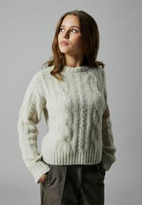 Pull en laine gris clair en tricot à torsades, avec un col rond côtelé et des motifs texturés. Le modèle porte un pantalon à carreaux.
