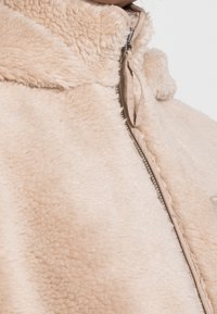 Beige fleecetrøje med en blød, tekstureret finish. Har en høj krave og en metal lynlås. Ingen synlige mønstre eller accenter.