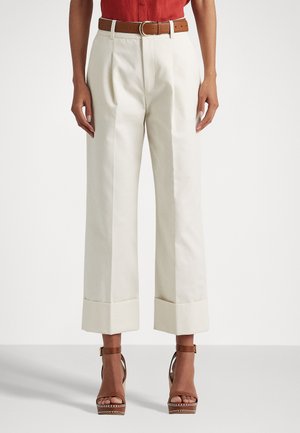 Pantalones anchos de color crema con cintura alta, con pliegues delanteros y dobladillos con puños. Combinados con un cinturón marrón y sandalias de tacón marrones con tiras.