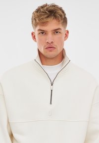 White quarter-zip pullover with a soft texture, featuring a silver zipper and a subtle logo detail on the chest.
