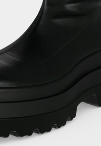 Bottine à plateforme en cuir noir avec semelle épaisse et robuste et talon bloc bas sur un fond clair uni.