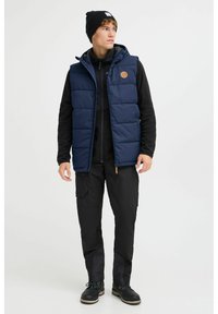 Navy Blouson-Weste mit Kapuze, Frontreißverschluss und Logo-Patch. Getragen über einem schwarzen Fleece-Oberteil und kombiniert mit schwarzen Cargo-Hosen und Stiefeln.