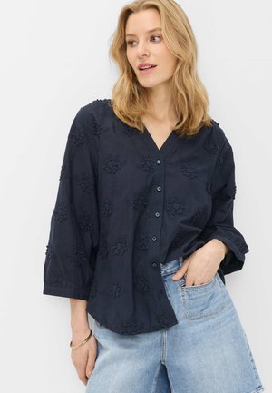 Frau mit blondem Haar, die eine marineblaue Bluse mit floralem Muster und Knöpfen sowie hellblaue Jeans trägt, mit der Hand in der Tasche.