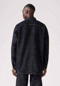 Donkerdenim overhemd met een ontspannen pasvorm, voorzien van een puntkraag, knoopmanchetten en zichtbare stikseldetails op de rug en zijnaden.