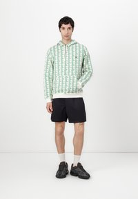 Felpa con cappuccio a righe verdi e bianche, con cordoncino, abbinata a pantaloni corti scuri. Il modello indossa sneakers grigie e calzini bianchi alla caviglia.