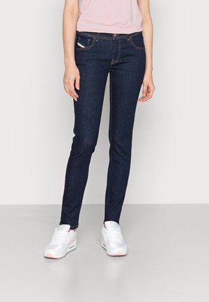 Personne portant un jean skinny bleu foncé, un haut rose clair et des baskets blanches, debout avec une posture détendue sur un sol gris clair uni.
