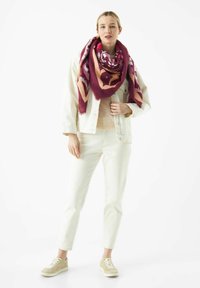 Oversized sjaal met patroon in bordeauxrood en roze stof, gecombineerd met een licht denim jack en witte broek, aangevuld met beige sneakers.