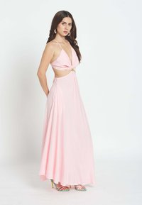 Mujer con cabello largo y oscuro, vestida con un vestido maxi sin mangas de color rosa claro con recorte en la cintura y sandalias de tacón alto coloridas, de pie de perfil.