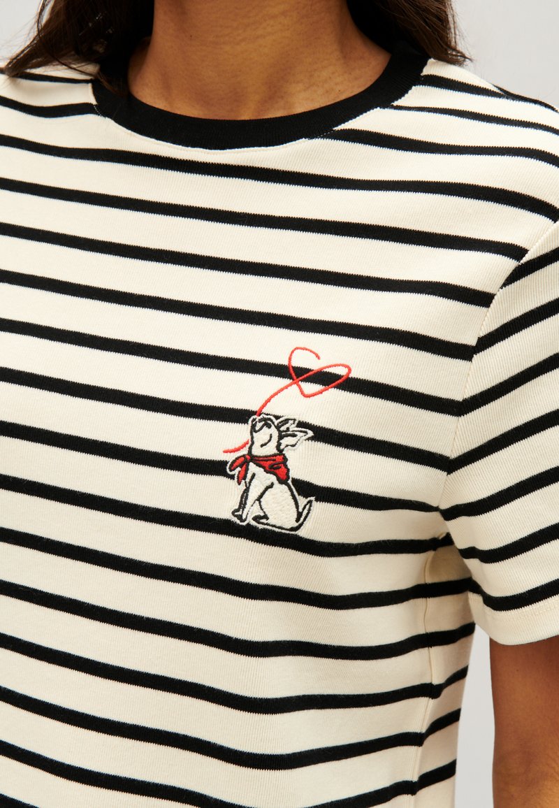 Koszulka w paski w kolorze kremowym i czarnym, z białym haftowanym psem w czerwonym szaliku i detalem w kształcie serca na klatce piersiowej. Krótkie rękawy, okrągły dekolt.