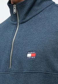 Pullover de fleece azul-marinho com fecho éclair e gola alta. Apresenta um pequeno logotipo bordado em vermelho, branco e azul na área do peito esquerdo.