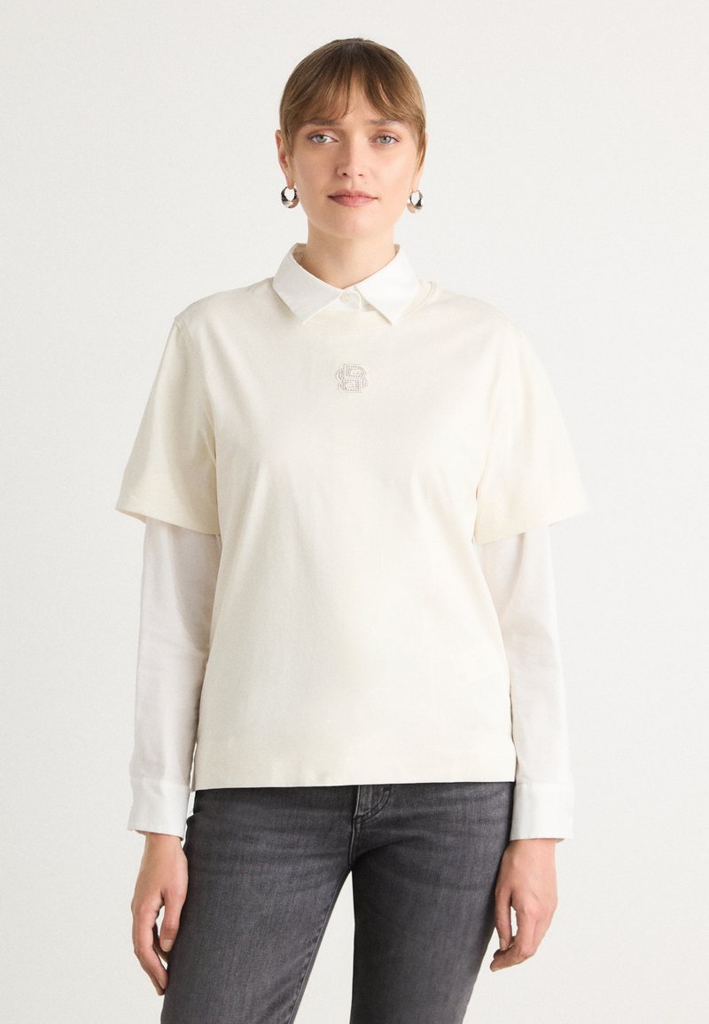 Camicia a maniche corte color crema con uno strato color bianco sotto il colletto. Presenta un piccolo logo di strass nell'area del petto.