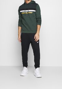 Zelená mikina s kapucí s černým, bílým a žlutým pruhovaným designem a logem na hrudi. Spárována s černými sportovními kalhotami a bílými sportovními botami.