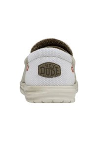 Baskets blanches à enfiler avec des accents beiges et une doublure texturée. Présente le logo "HEY DUDE" sur un patch en caoutchouc à l'arrière.