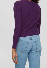 Lila gerippter Strickpullover mit langen Ärmeln, getragen mit hellblauen Jeans. Der Pullover hat eine glatte Textur und einen figurbetonten Schnitt.