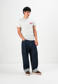 Homme debout avec les mains dans les poches, portant un t-shirt blanc Tommy Jeans, un jean large bleu foncé retroussé, et des baskets marron sur fond blanc.
