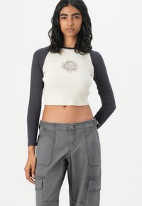 Cropped long-sleeve shirt with a light cream body and dark navy sleeves, featuring a sun graphic in beige at the center. Paired with gray cargo pants.