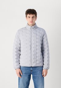 Lichtgrijze pufferjack met hoge kraag, vierkant doorstikte ontwerp, ritssluiting en elastische manchetten, gecombineerd met blauwe denim jeans.